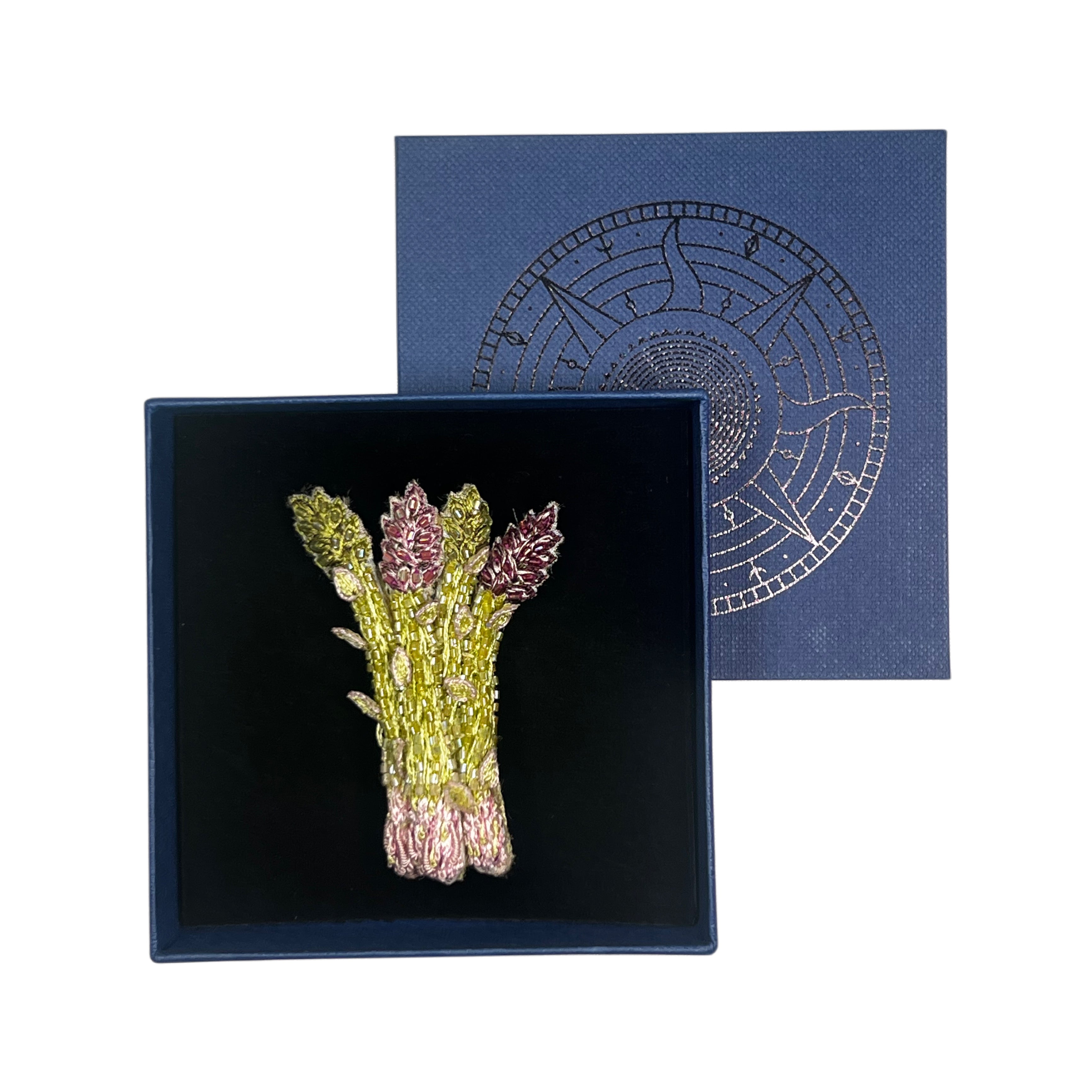 An asparagus brooch featuring green and pink metallic detailing, presented in a black velvet-lined navy blue box with its matching navy blue lid shown behind it.