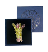 An asparagus brooch featuring green and pink metallic detailing, presented in a black velvet-lined navy blue box with its matching navy blue lid shown behind it.